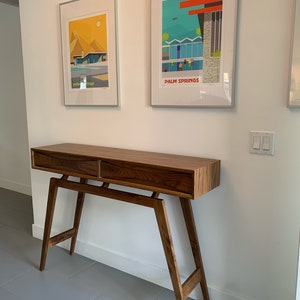 May include: A dark brown wooden console table with two drawers and a mid-century modern design. The table has a simple, clean design with angled legs and a rectangular top. The table is positioned in front of a white wall with two framed prints of Palm Springs, California.