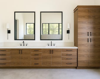 May include: A double vanity bathroom cabinet with a wood grain finish, featuring two white sinks, black hardware, and two rectangular mirrors with black frames. A tall matching wood storage cabinet stands to the right, flanked by two wall sconces.