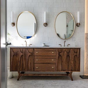 K&ouml;nnte beinhalten: Ein moderner Badezimmer-Waschtisch mit Nussbaumholz-Finish und goldenen Beschl&auml;gen. Der Waschtisch hat drei Schubladen und zwei Waschbecken mit runden Spiegeln &uuml;ber jedem Waschbecken.