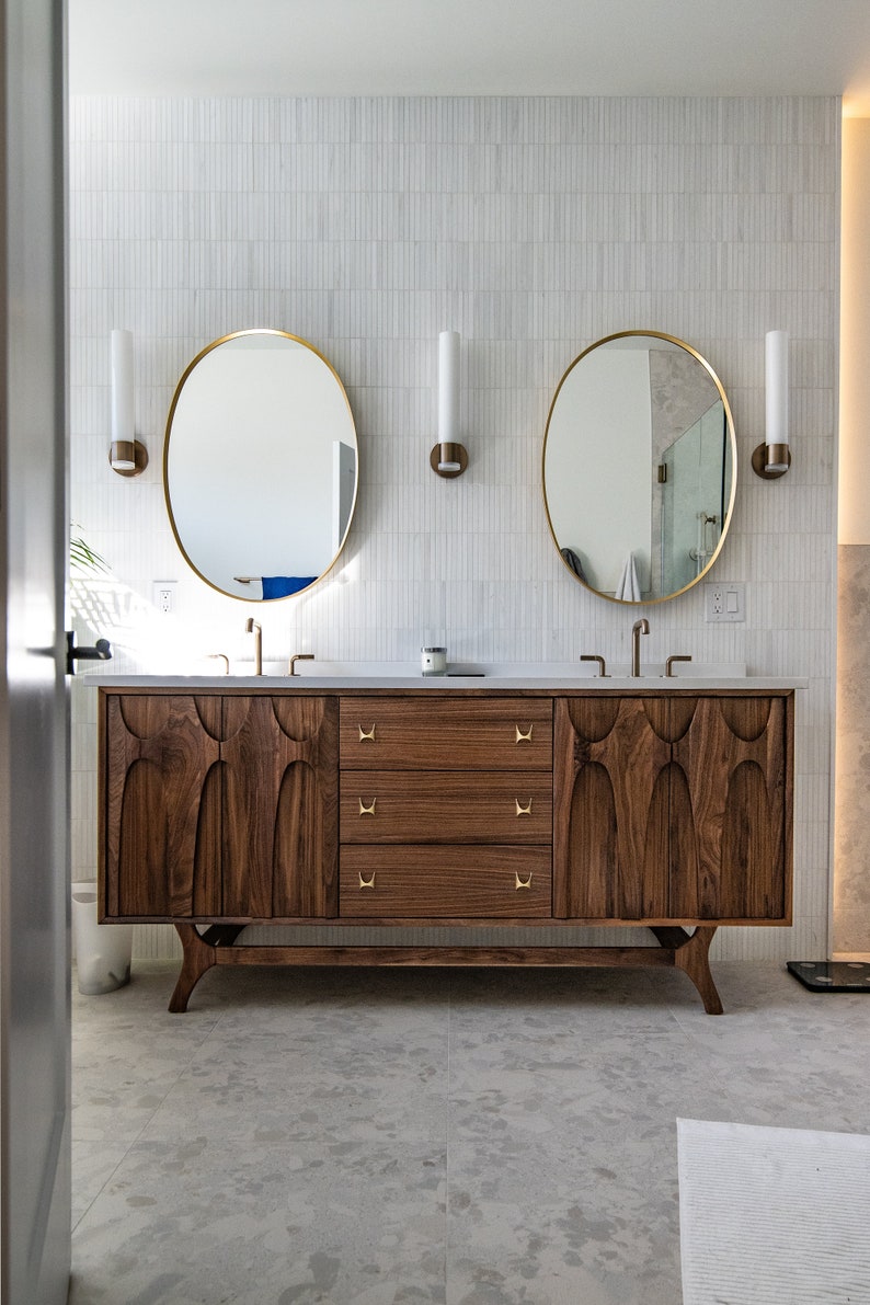 K&ouml;nnte beinhalten: Ein moderner Badezimmer-Waschtisch mit Nussbaumholz-Finish und goldenen Beschl&auml;gen. Der Waschtisch hat drei Schubladen und zwei Waschbecken mit runden Spiegeln &uuml;ber jedem Waschbecken.