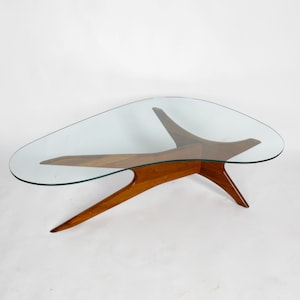 May include: A mid-century modern coffee table with a glass top and a unique, three-legged wooden base. The base is made of dark wood and has a distinctive, organic shape.