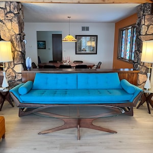 May include: A mid-century modern living room featuring a turquoise sofa with a wooden frame and a glass coffee table. Two white lamps with beige shades flank the sofa. The room has stone columns and an orange ottoman.