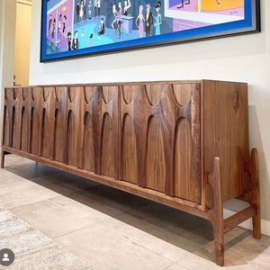 May include: A long, dark wood cabinet with a unique, carved design. The cabinet has multiple doors and sits on tall, slender legs.