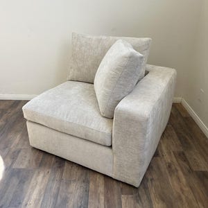 May include: A white, fabric-covered corner sectional sofa with a matching pillow. The sofa has a tufted design and a square shape.