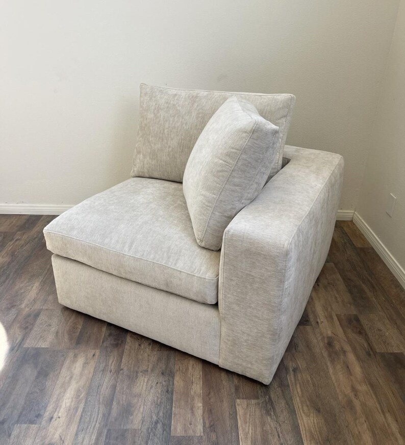 May include: A white, fabric-covered corner sectional sofa with a matching pillow. The sofa has a tufted design and a square shape.