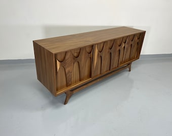 May include: A mid-century modern walnut wood sideboard with sliding doors featuring a unique carved design. The sideboard has a rich brown finish and sits on four angled legs.