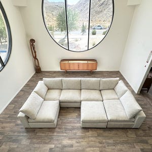 May include: A large, off-white sectional sofa with multiple cushions and a modular design. The sofa is positioned in a room with hardwood floors and a large circular window.