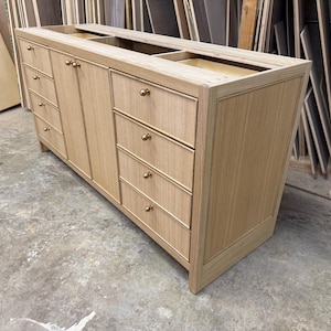 May include: A light-colored wooden bathroom vanity with multiple drawers and cabinet doors. The vanity has gold-colored knobs and is unfinished, showing the natural wood grain. The design includes two recessed areas on the top, likely for sinks.