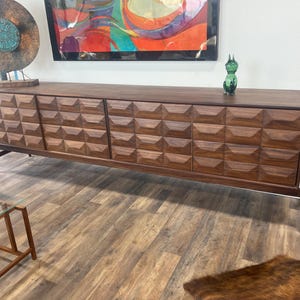 May include: A mid-century modern walnut credenza with geometric carved door fronts. The long, low cabinet has four doors and sits on angled legs. A green glass figurine sits on top. The credenza is a stylish storage solution.