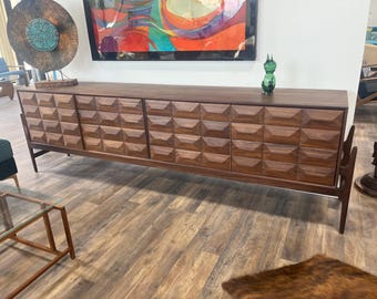 May include: A mid-century modern walnut credenza with geometric carved door fronts. The long, low cabinet has four doors and sits on angled legs. A green glass figurine sits on top. The credenza is a stylish storage solution.