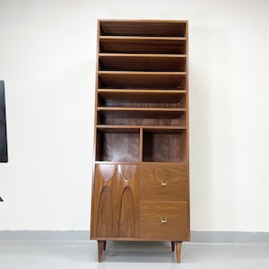 Pu&ograve; includere: Una libreria alta in legno in stile moderno di met&agrave; secolo con pi&ugrave; ripiani e un mobile inferiore con due cassetti. La libreria &egrave; realizzata in legno dai toni caldi e presenta un design intagliato unico sulle ante dell'armadio. I cassetti hanno maniglie color oro.