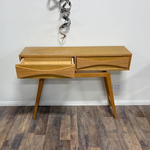 May include: A mid-century modern wooden console table with two drawers, one open. The table has angled legs and a light brown finish. The drawer fronts have a curved design. The table is set against a white wall.