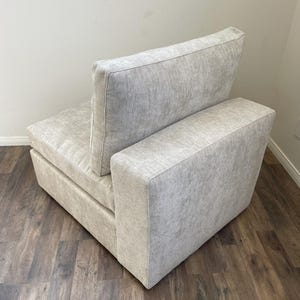 May include: A light gray fabric armchair with a single armrest and a back cushion. The chair is sitting on a wood floor.