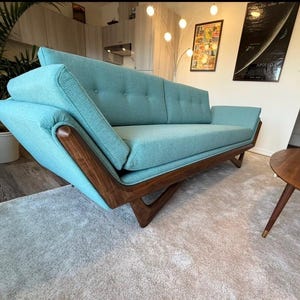 May include: A mid-century modern style turquoise sofa with a wooden frame. The sofa has a unique angled armrest design and button tufting on the back cushions. The sofa is set on a light gray rug.