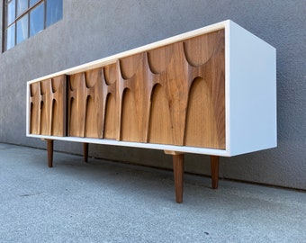 May include: A mid-century modern sideboard with a white painted exterior and wood doors with a unique carved design. The sideboard has four legs and is made of wood.