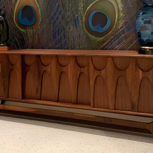 May include: A long, dark wood sideboard with a unique, carved design on the doors. The sideboard has a sleek, mid-century modern style and sits on four angled legs.