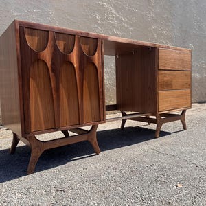 May include: A mid-century modern walnut wood desk with a unique carved design on the front doors. The desk has three drawers on the right side and a open shelf on the left side. The desk is supported by four angled legs.