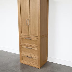 May include: Tall wooden cabinet with two doors at the top and three drawers below. The cabinet is made of light-colored wood and has silver-toned handles. It stands against a white wall.