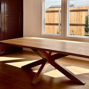 May include: A large, rectangular wooden dining table with a unique, angled base. The table is made of light-colored wood and is positioned in a room with hardwood flooring and natural light from a nearby window. The table's dimensions are approximately 96 inches long and 40 inches wide.