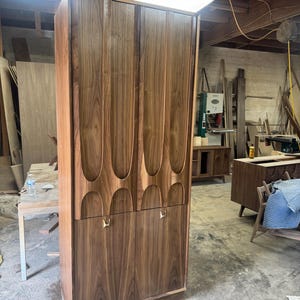 May include: A tall, mid-century modern wooden cabinet with a dark walnut finish. The upper doors feature a series of carved, elongated oval shapes. The lower doors have brass-colored handles. The cabinet is in a workshop setting.