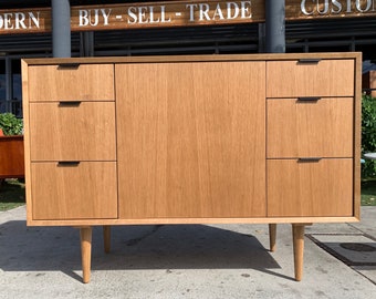 May include: A light wood dresser with three drawers on each side and a cabinet door in the center. The dresser has four tapered legs and black drawer pulls.