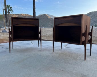 May include: Two mid-century modern nightstands made of dark wood. Each nightstand has a drawer with a curved design, an open shelf, and unique side supports. The nightstands are set on thin, tapered legs.
