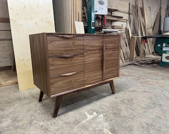 May include: A mid-century modern style wooden cabinet with three drawers and a door. The cabinet is made of dark brown wood and has a minimalist design with angled legs. The drawers have curved handles.