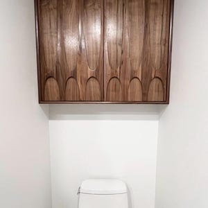 May include: A dark wood cabinet with carved vertical and arched designs is mounted above a white toilet. The cabinet has a rectangular frame and two doors. The bathroom setting is simple and modern.