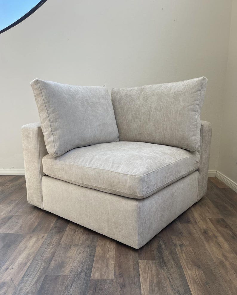 May include: A light gray corner sectional sofa with two plush cushions. The sofa has a tufted fabric upholstery and a square shape.