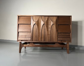May include: A mid-century modern walnut dresser with a unique carved design on the doors. The dresser has four drawers on each side and sits on a set of four legs.
