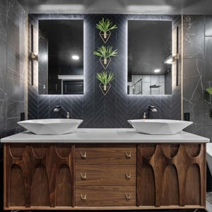 K&ouml;nnte beinhalten: Ein modernes Badezimmer-Waschbecken mit einer dunkelgrauen Fliesenwand, zwei wei&szlig;en Porzellanwaschbecken und einem Schrank aus Holz mit einem einzigartigen geschnitzten Design. Das Waschbecken hat drei Schubladen und einen gro&szlig;en Spiegel &uuml;ber den Waschbecken.