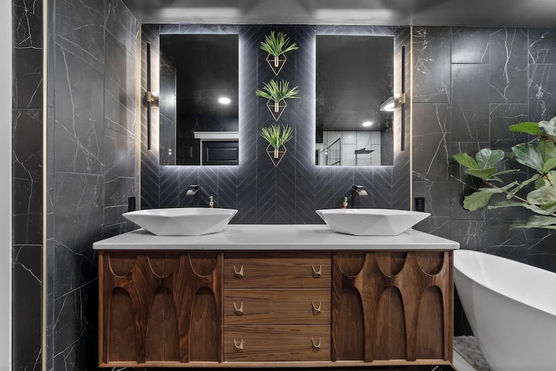 K&ouml;nnte beinhalten: Ein modernes Badezimmer-Waschbecken mit einer dunkelgrauen Fliesenwand, zwei wei&szlig;en Porzellanwaschbecken und einem Schrank aus Holz mit einem einzigartigen geschnitzten Design. Das Waschbecken hat drei Schubladen und einen gro&szlig;en Spiegel &uuml;ber den Waschbecken.