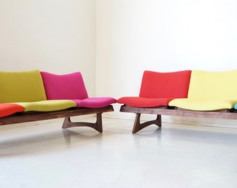 May include: A set of two mid-century modern modular sofas with a walnut wood frame and colorful fabric upholstery. The sofas are arranged in a row with the cushions in a pattern of red, yellow, pink, and blue.