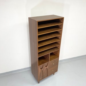 Pu&ograve; includere: Un mobile contenitore alto in legno in stile moderno della met&agrave; del secolo, con pi&ugrave; ripiani aperti, due piccoli cassetti e dettagli decorativi intagliati nella sezione inferiore. Il mobile &egrave; di colore marrone caldo e poggia su gambe corte e angolate.