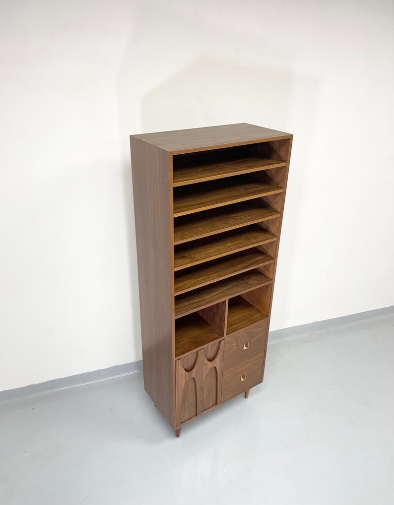 Pu&ograve; includere: Un mobile contenitore alto in legno in stile moderno della met&agrave; del secolo, con pi&ugrave; ripiani aperti, due piccoli cassetti e dettagli decorativi intagliati nella sezione inferiore. Il mobile &egrave; di colore marrone caldo e poggia su gambe corte e angolate.