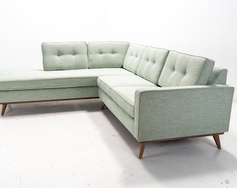 May include: A light green sectional sofa with tufted cushions and wooden legs. The sofa is in a modern style and has a clean, minimalist design.