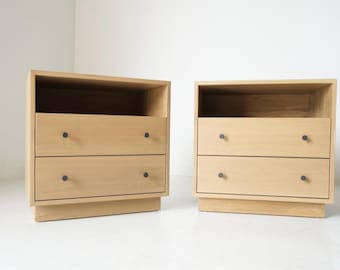 May include: Two matching wooden nightstands with two drawers each and a top shelf. The nightstands are light brown with dark gray knobs. The nightstands are on a white floor and against a white wall.