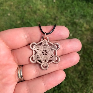May include: A rose gold pendant necklace featuring a geometric design with a black cord. The pendant is a detailed, intricate design.