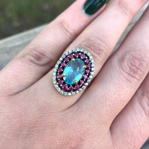 Puede incluir: Un anillo de plata con una gran piedra azul ovalada rodeada de piedras rojas y blancas más pequeñas.