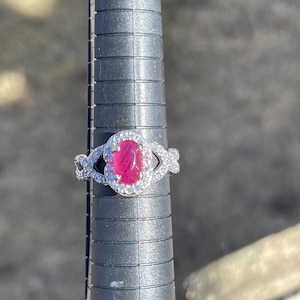 Puede incluir: Un anillo de plata con una gran piedra de rubí ovalada rodeada de diamantes más pequeños. El anillo está en un medidor de tamaño de anillo.