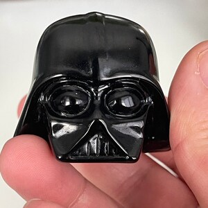 May include: A black plastic figurine of Darth Vader from Star Wars. The figurine has a detailed face with a black helmet and a black mask.