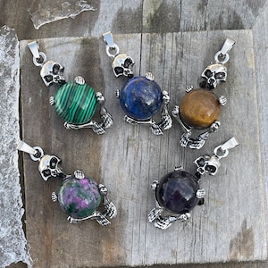 Pode incluir: Cinco pingentes de pedras preciosas com desenhos de caveiras e mãos de esqueleto. As pedras preciosas são verdes, azuis, castanhas, rosas e roxas. Os pingentes são exibidos em uma superfície de madeira envelhecida.