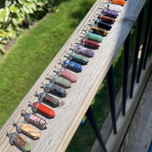 May include: A collection of 20 colorful crystal point pendants with silver caps. The pendants are arranged in a row on a wooden surface.