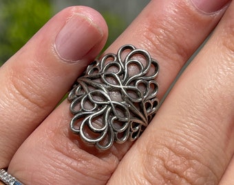 Puede incluir: Un anillo de filigrana de plata con un diseño delicado e intrincado. El anillo está hecho de un alambre delgado y retorcido que forma una serie de círculos y bucles superpuestos.