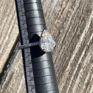 May include: A pear-shaped diamond engagement ring with a pave setting on a silver band. The ring is being sized on a ring sizer that shows the ring size is 10.