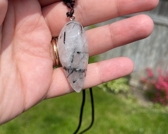 May include: A clear quartz pendant with black tourmaline inclusions, hanging from a brown cord with black and white beads.