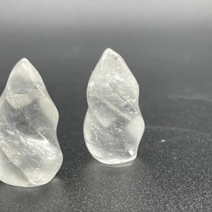 Puede incluir: Dos cristales de cuarzo transparentes, cada uno tallado en forma de espiral, se muestran sobre una superficie gris oscuro. Los cristales son translúcidos y tienen un acabado liso y pulido.
