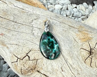 May include: A teardrop-shaped pendant with a silver bail. The pendant features a polished, dark green stone with lighter green and white marbling. The pendant is displayed on a piece of weathered driftwood, with small white stones in the background.