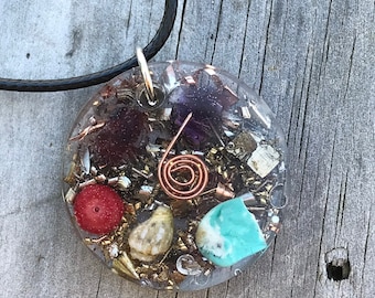 Op de afbeelding: Een ronde, transparante hars hanger ketting met een zwart koord. De hanger bevat verschillende kleine stukjes gekleurde stenen, metaal en een koperen draadspiraal.