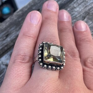 Op de afbeelding: Een zilveren ring met een grote, vierkante, lichtgroene edelsteen gezet in een geperlde zetting.